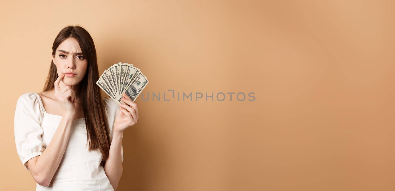 Pensive serious girl holding money and thinking, looking thoughtful at camera and frowning, deciding what to buy with dollar bills, beige background.