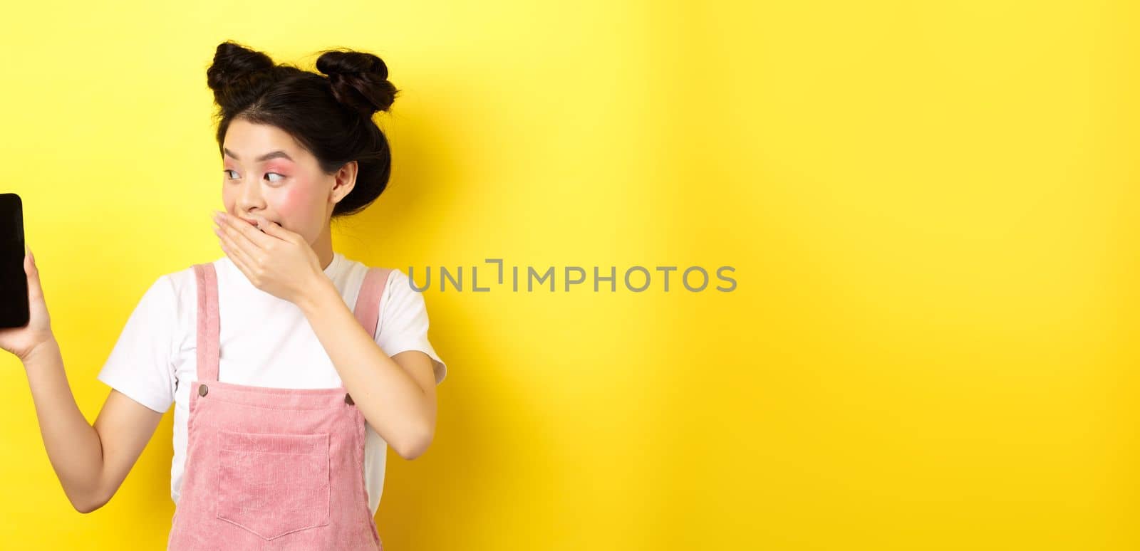 Online shopping concept. Silly japanese girl with beauty makeup, cover mouth with hand laughing and showing empty smartphone screen, show funny thing on phone, yellow background.