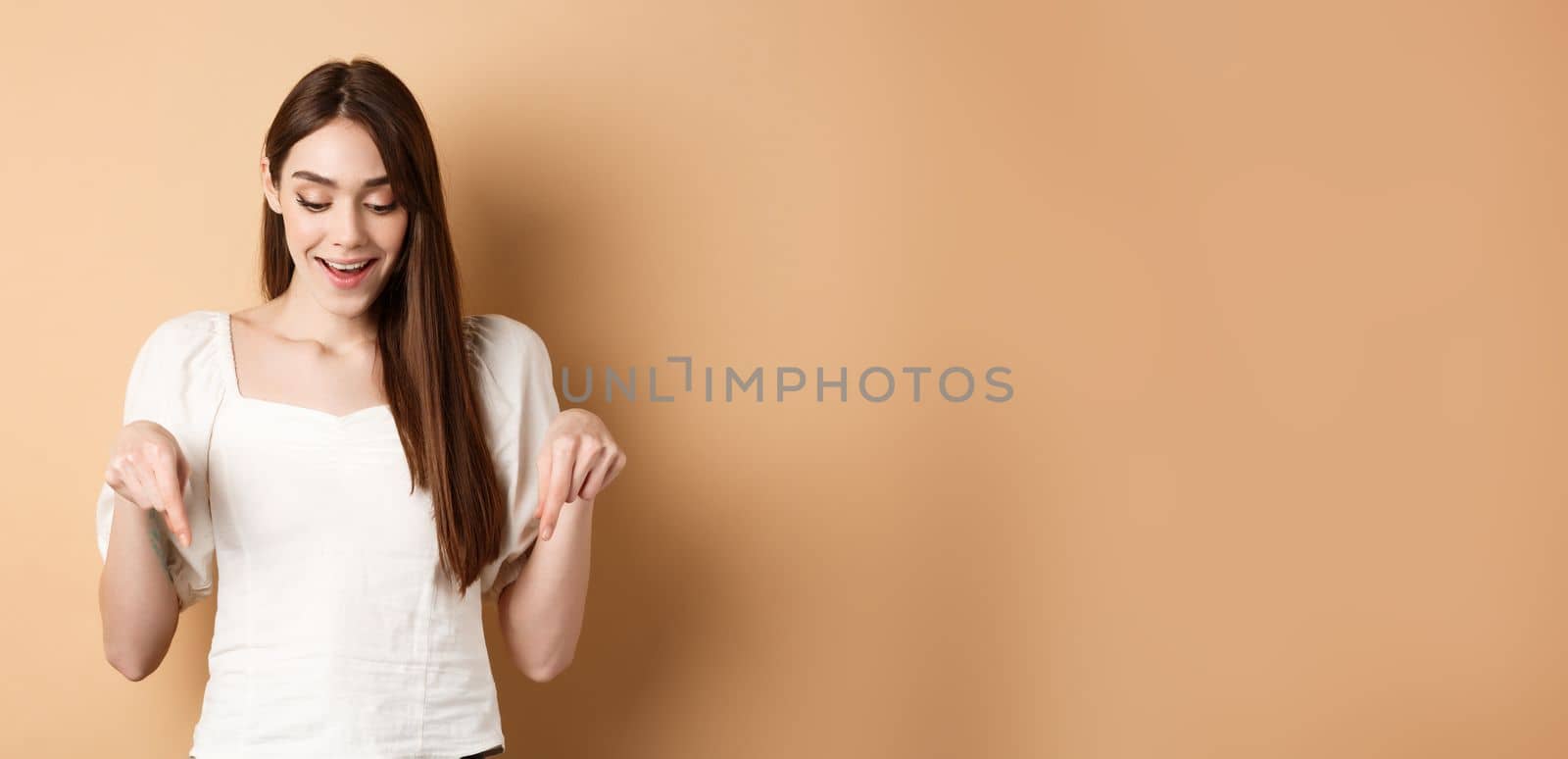 Surprised cute girl say wow, smiling amazed and pointing fingers down, showing advertisement, standing on beige background.