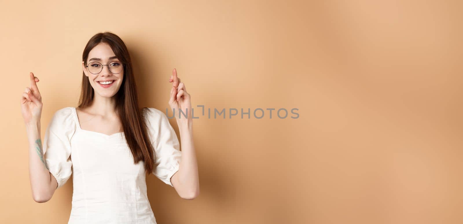 Hopeful smiling girl making wish, cross fingers for good luck and looking happy, praying for dream to come true, standing on beige background by Benzoix