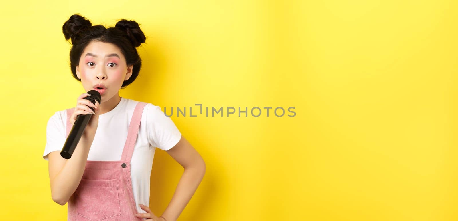 Cute asian teen girl with bright makeup, singing in microphone karaoke, standing against yellow background.