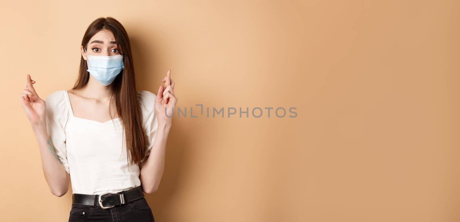 Covid-19 and lifestyle concept. Hopeful girl in face mask praying with fingers crossed, looking with hope at camera, standing on beige background.