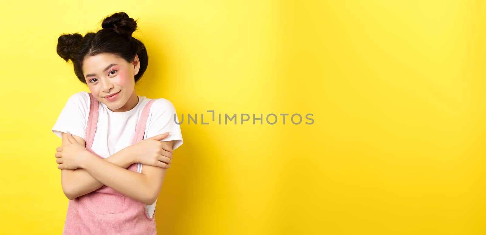 Summer lifestyle concept. Cute asian beauty girl with makeup, hugging herself and smiling romantic, standing tender on yellow background.
