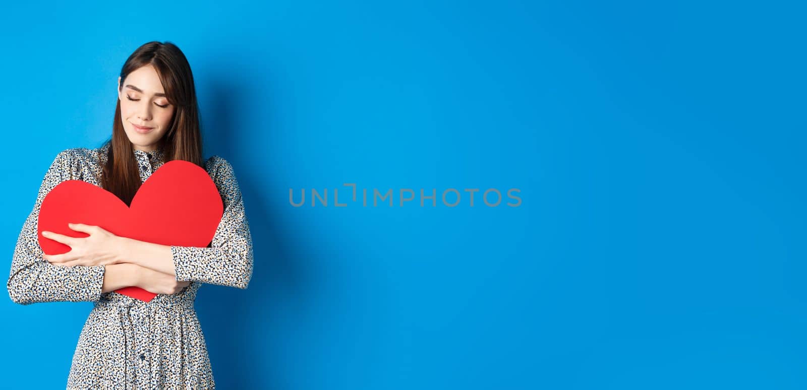 Valentines day. Romantinc girl in dress hugging big red heart cutout, close eyes and smile with dreamy face, imaging sensual date, standing on blue background.