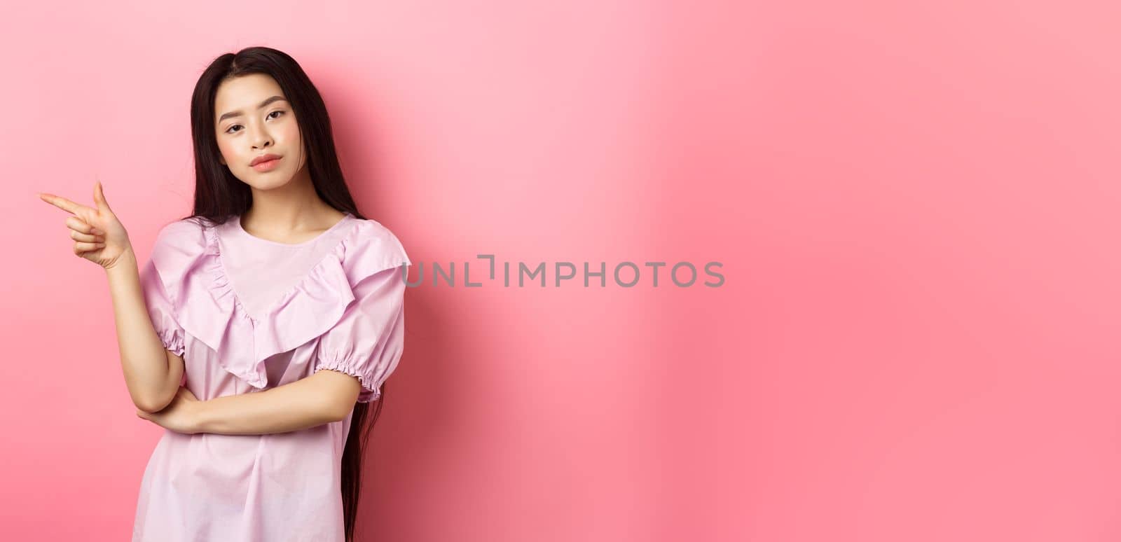 Young smug asian girl looking cool and pointing finger left at logo, advertising product on pink romantic background.