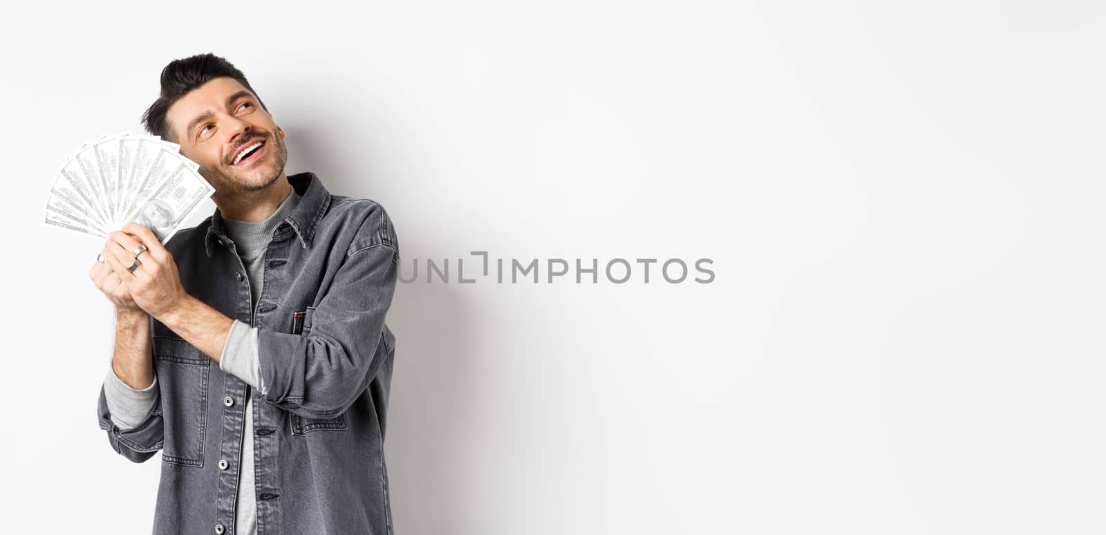 Happy rich guy hugging dollar bills and daydreaming, thinking of shopping with money, standing on white background.