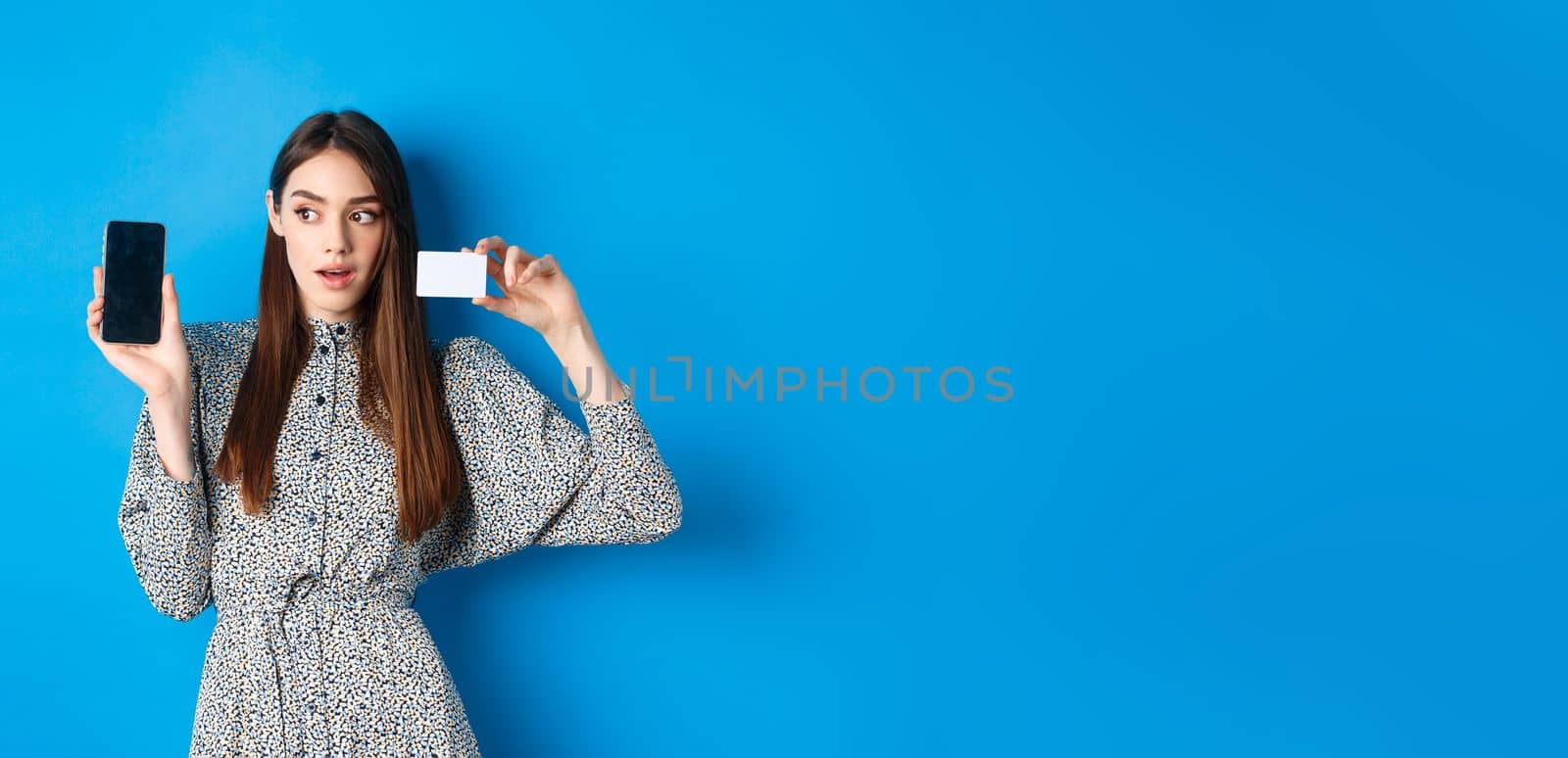 Online shopping. Pensive lady in dress showing empty phone screen and plastic credit card, order in internet, blue background.