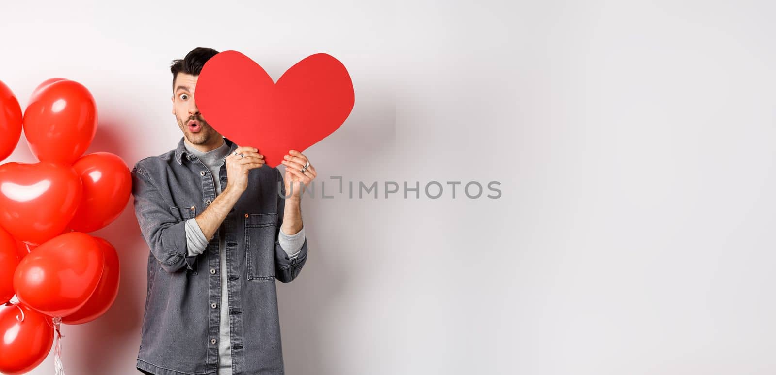 Cute young man hiding face behind big Valentine heart card, secret admirer looking at camera with amazement, saying wow, standing near romantic balloons on white background by Benzoix