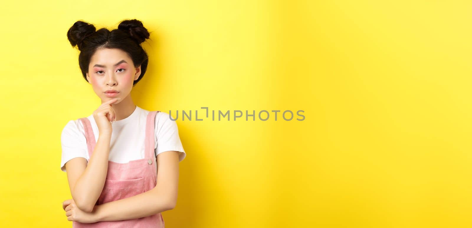 Suspicious asian girl looking at camera and thinking, stare with disbelief, standing with makeup on yellow background.