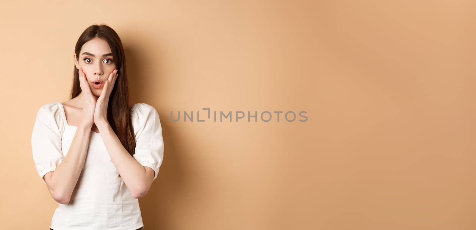 Wow unbelievable. Impressed young woman gasping and looking astonished by news, staring at camera, touching face in awe, standing on beige background by Benzoix