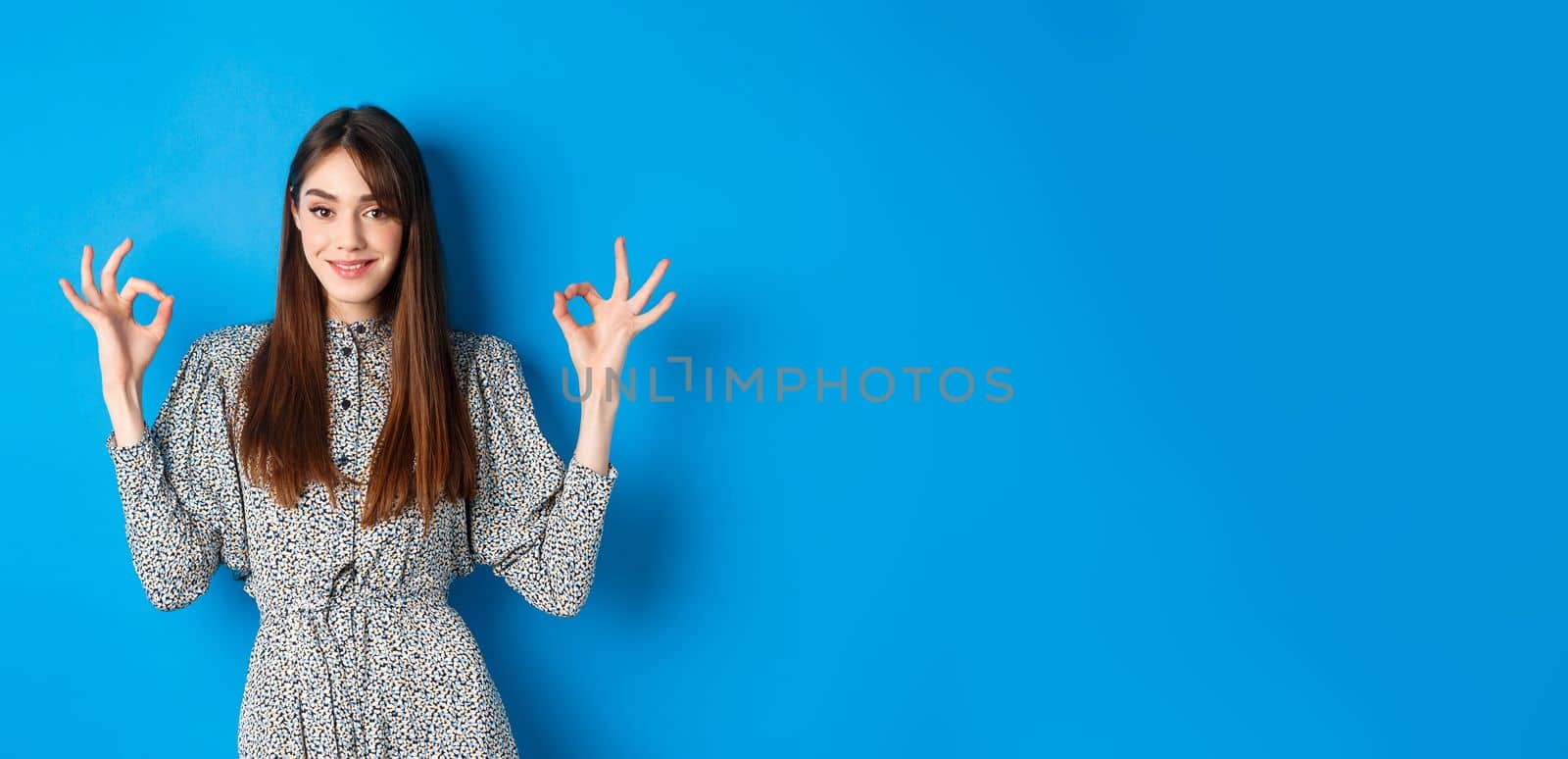 Confident adult woman in dress with natural hair color, showing okay signs and smiling, like and approve something good, standing on blue background.