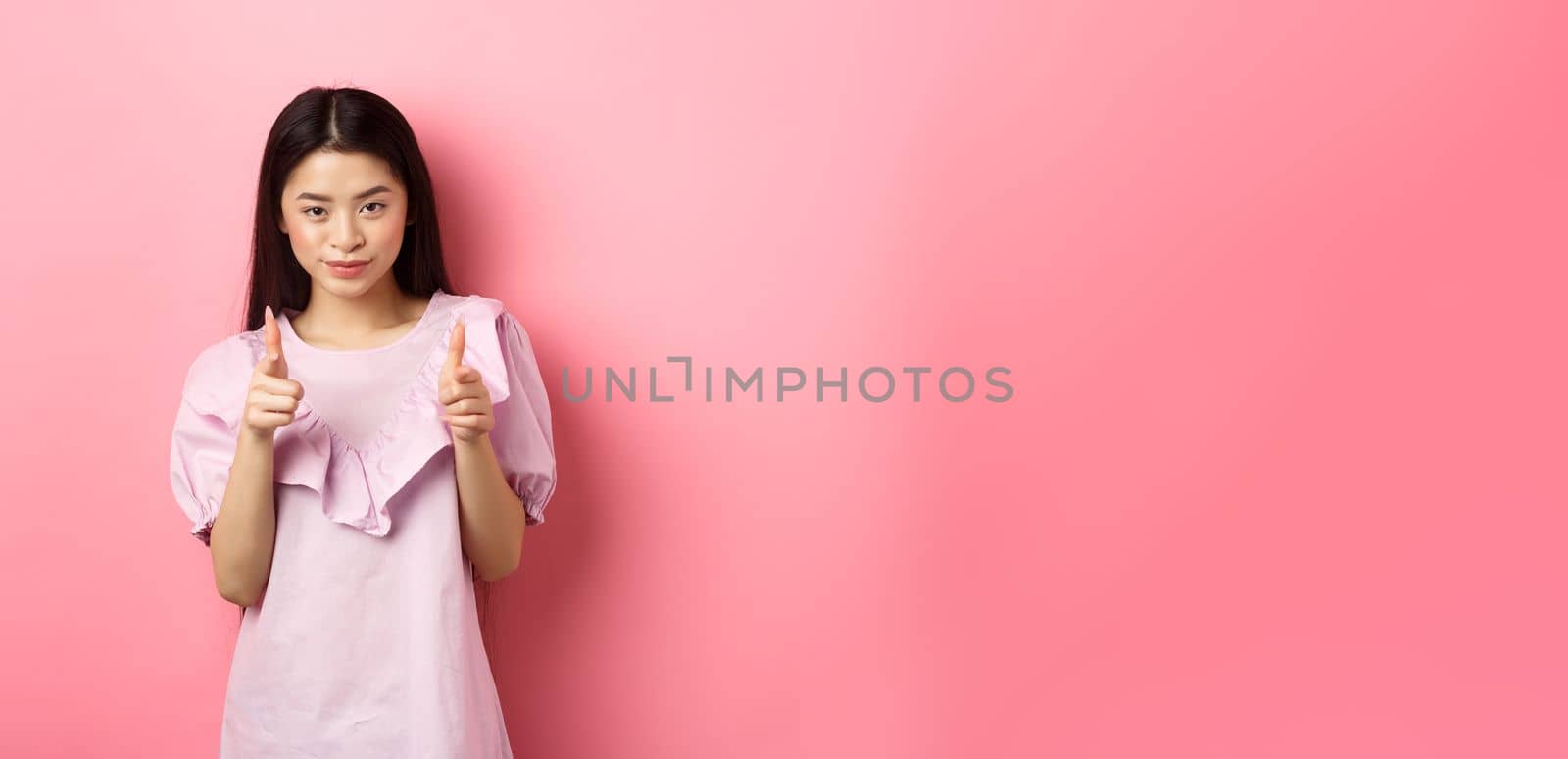 Sassy asian woman pointing at camera, smiling and inviting you, beckon or praise person, standing on pink background.