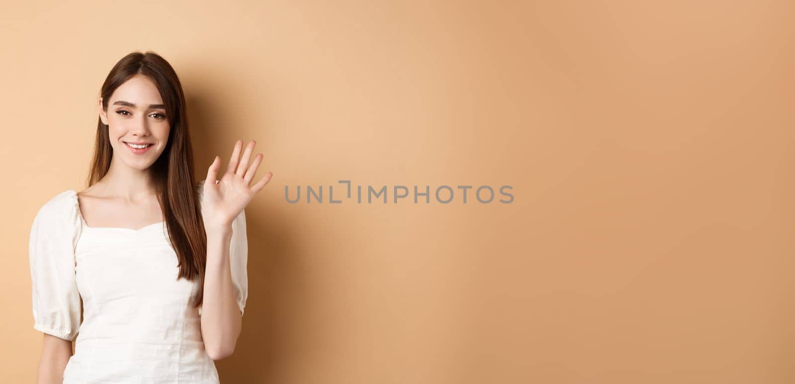 Friendly smiling woman say hello, waiving hand to greet you, standing cheerful on beige background.