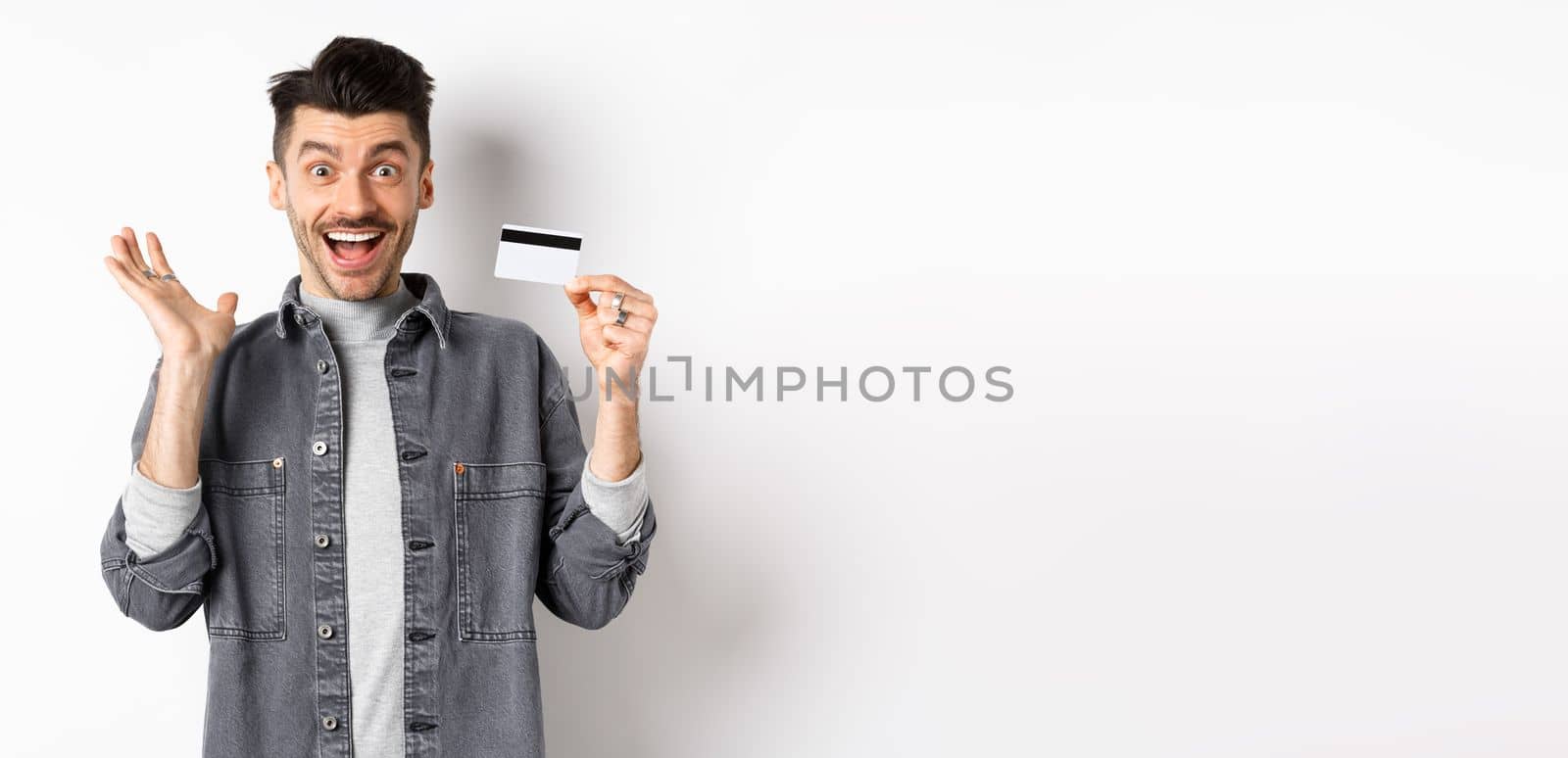 Excited man scream from joy and showing plastic credit card, rejoicing on shopping, standing against white background.