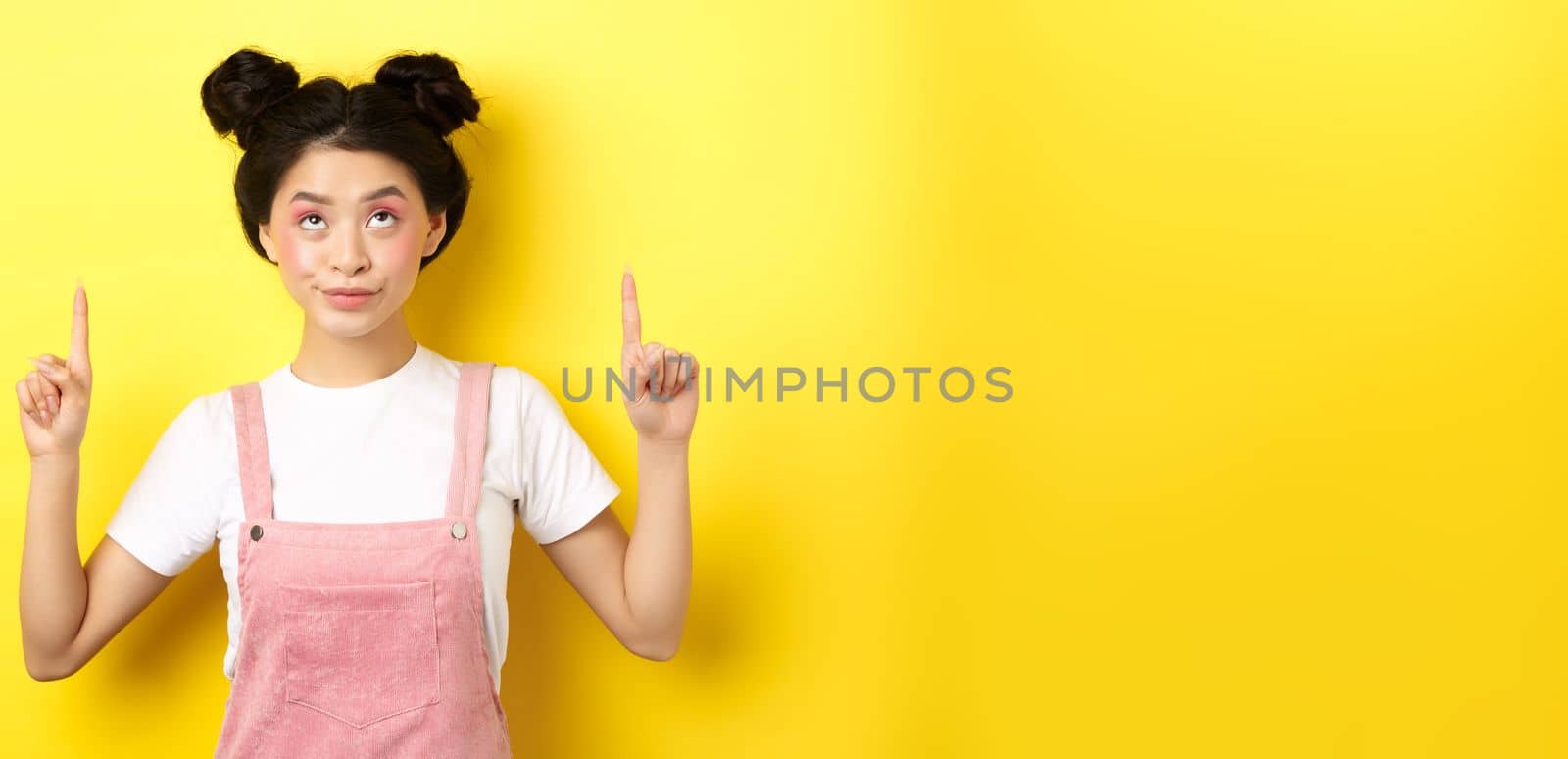 Glamour asian girl with beauty makeup, pointing fingers up, showing top advertisement, standing on yellow background by Benzoix