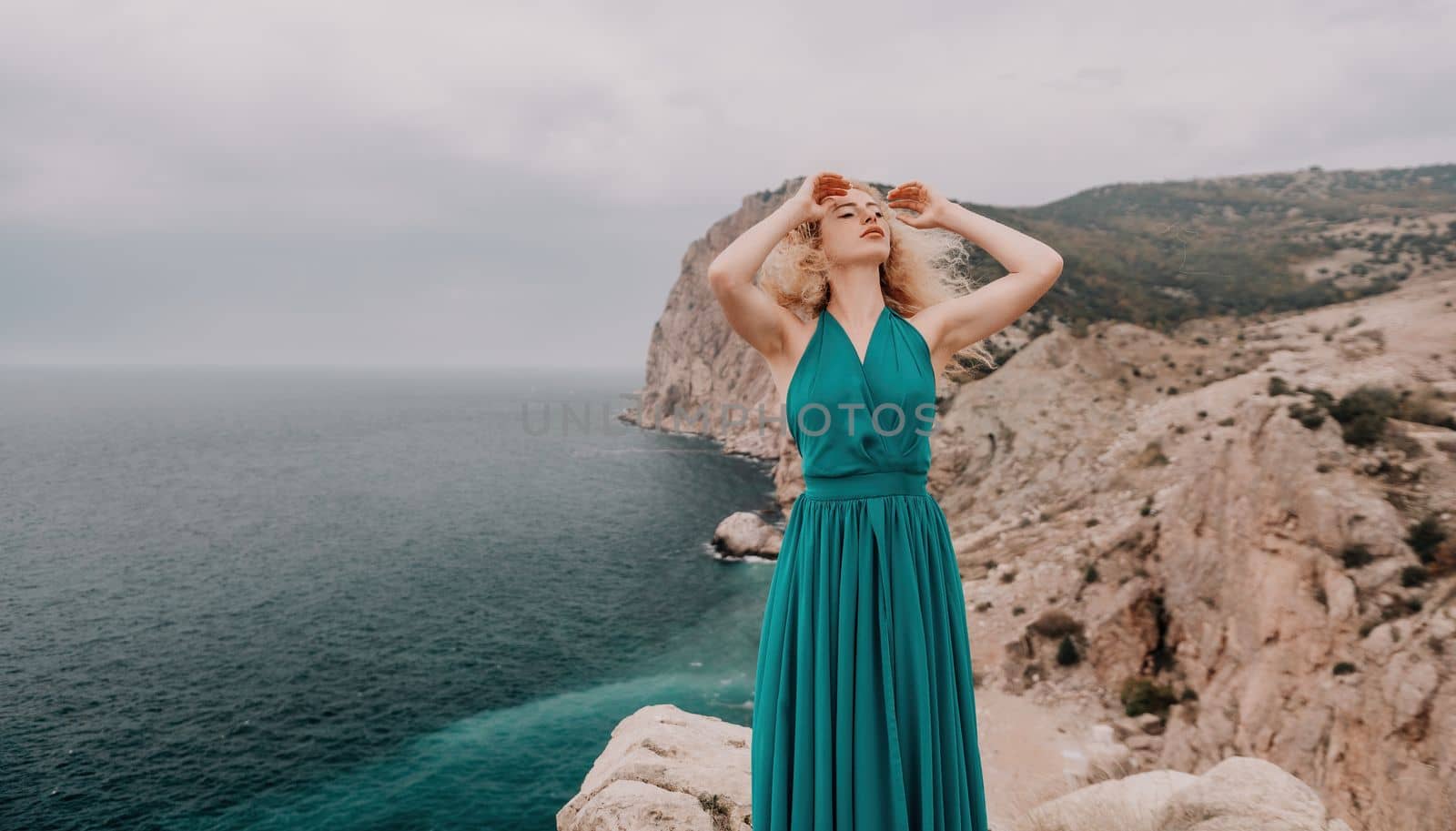 Side view a Young beautiful sensual woman in a mint long dress posing on a volcanic rock high above the sea during sunset. Girl on the nature on overcast sky background. Fashion photo