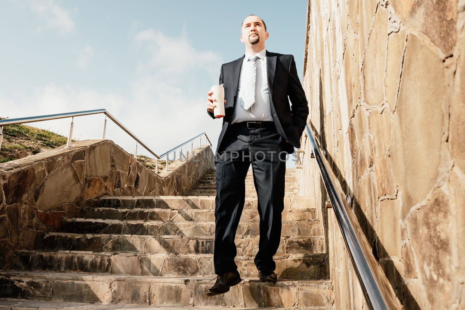 Confident middle age businessman with briefcase walking downstairs. Close-up of businessman wearing blue suit holding bag and coffee cup walking down the stairs by panophotograph