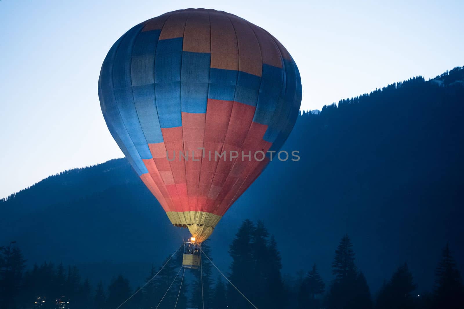 Hot air balloon with fire heating air in wicker basket with himalaya mountains in background showing this adventure in kullu manali valley India