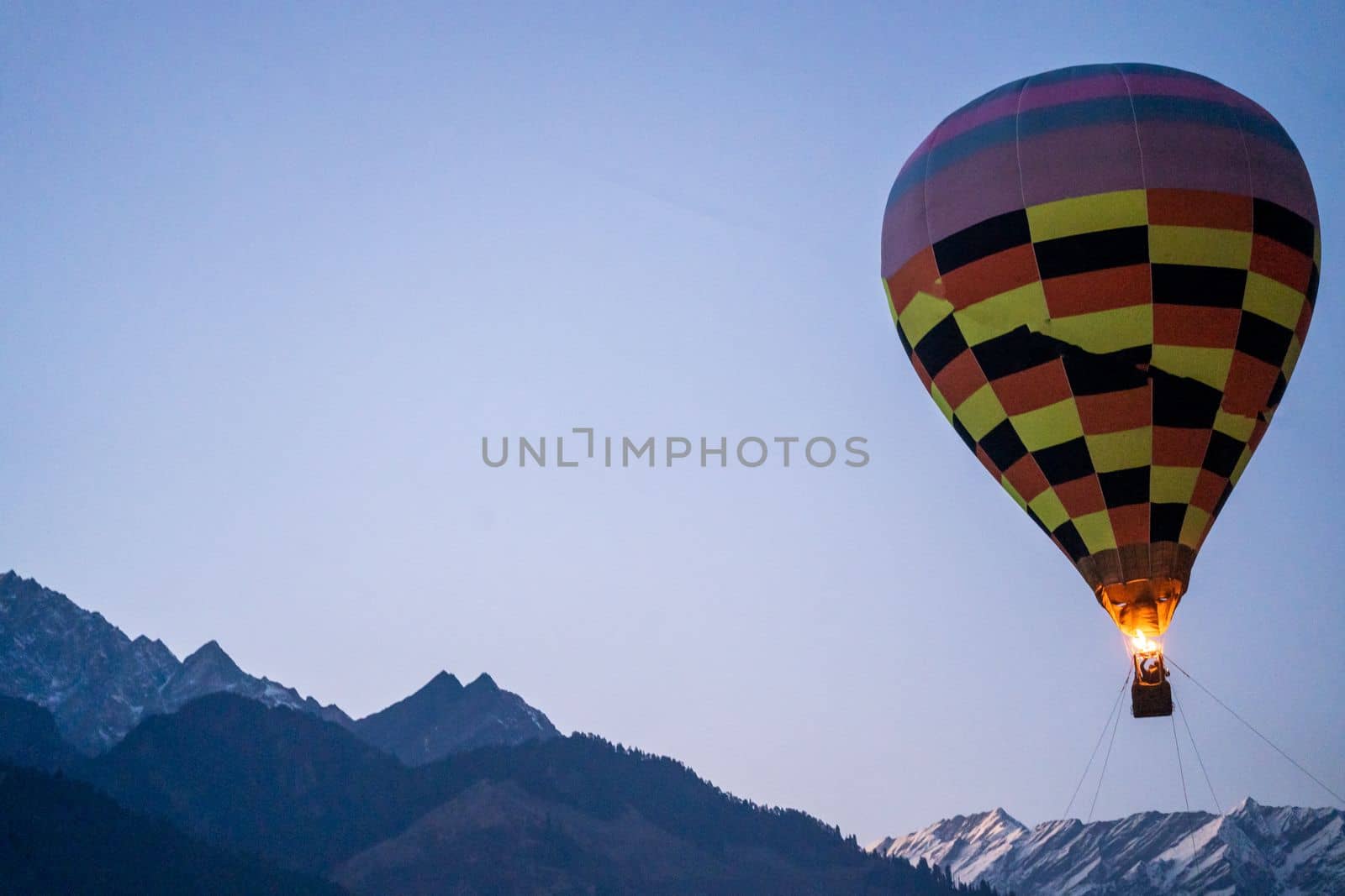 Hot air balloon with fire heating air in wicker basket with himalaya mountains in background showing this adventure in kullu manali valley India