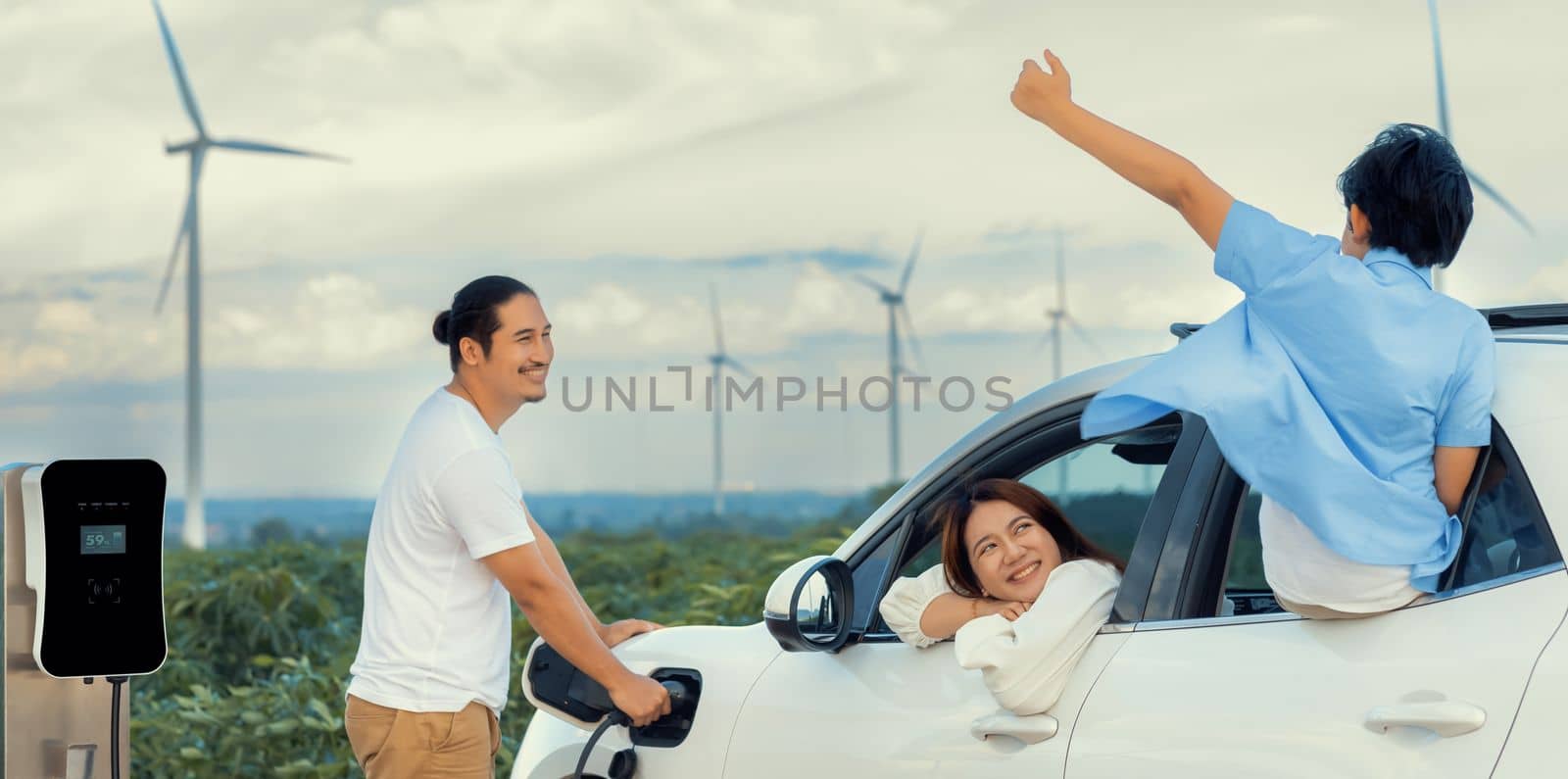 Concept of progressive happy family enjoying their time at wind farm with electric vehicle. Electric vehicle driven by clean renewable energy from wind turbine generator for charging station.