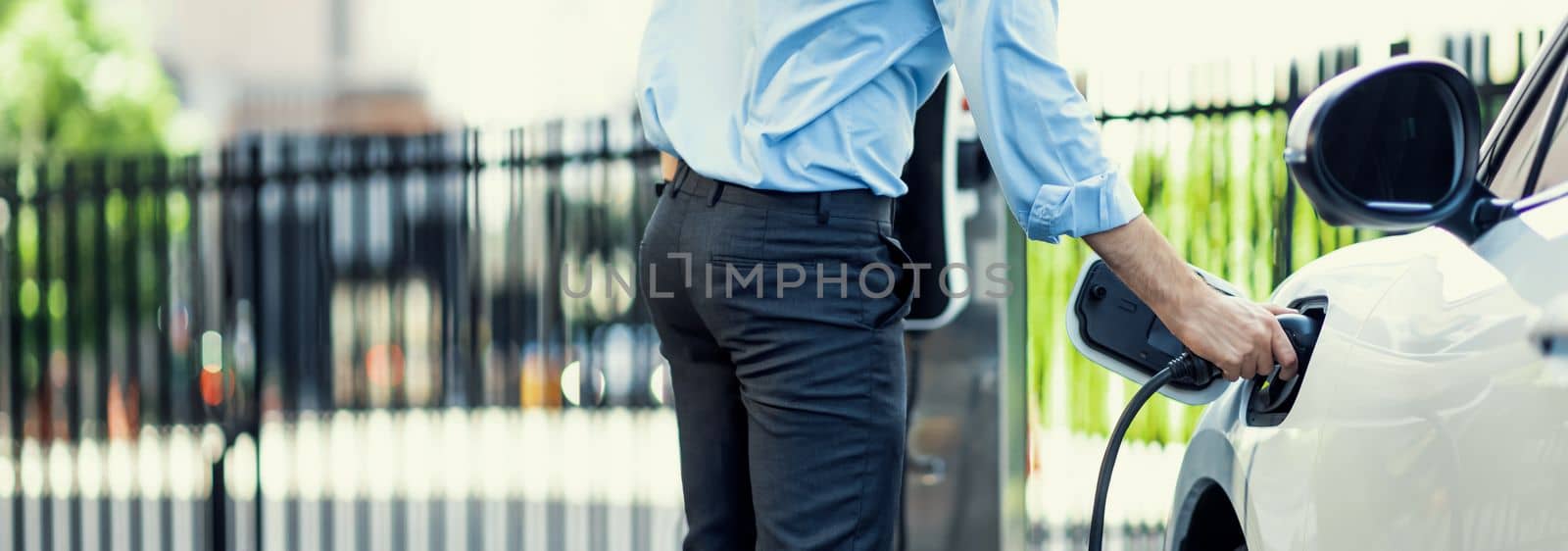 Closeup progressive man holding EV charger plug from public charging station for electric vehicle with background of residential building as concept eco-friendly sustainability energy car concept.