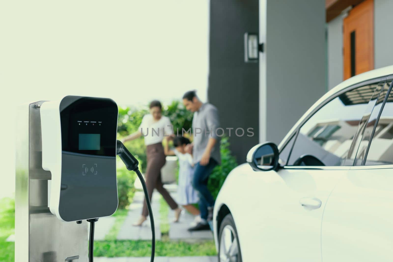 Focus closeup electric vehicle recharging battery from home electric charging station with blurred family in background. Renewable clean energy car for progressive eco awareness lifestyle concept.