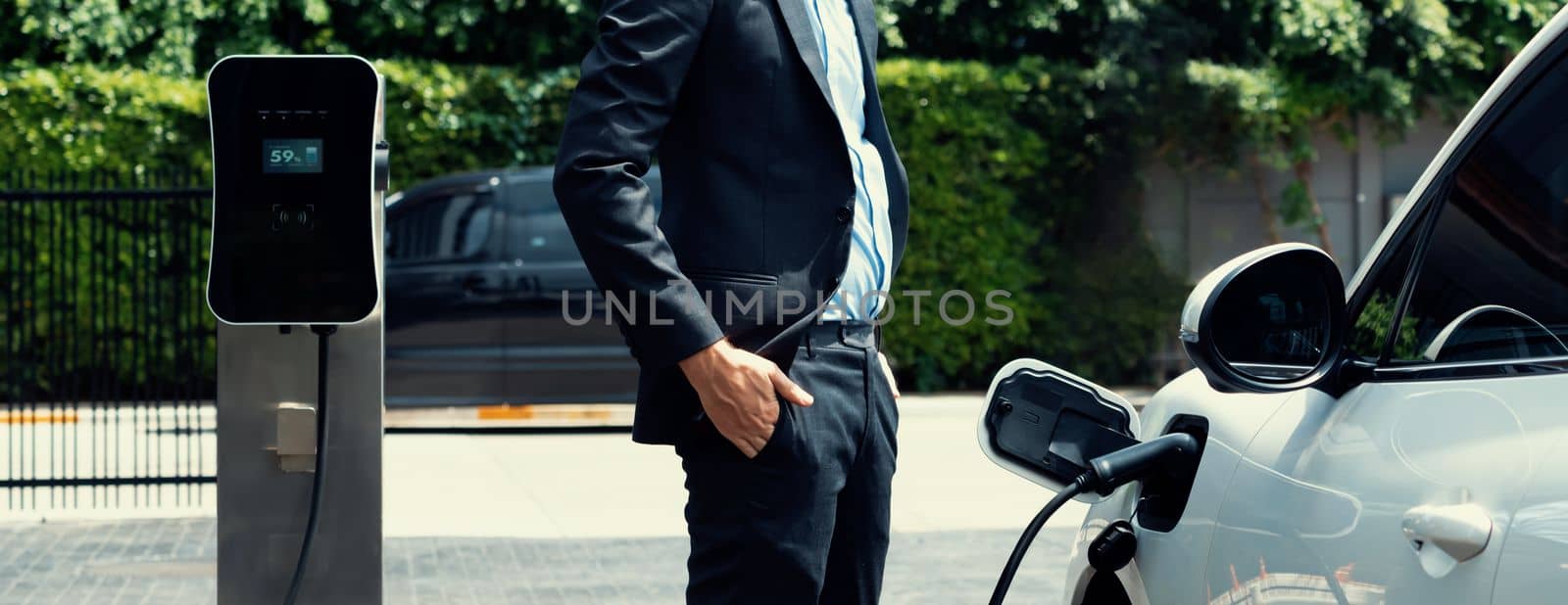 Closeup progressive suit-clad businessman with his electric vehicle recharge his car on public charging station in modern city with power cable plug and renewable energy-powered electric vehicle.