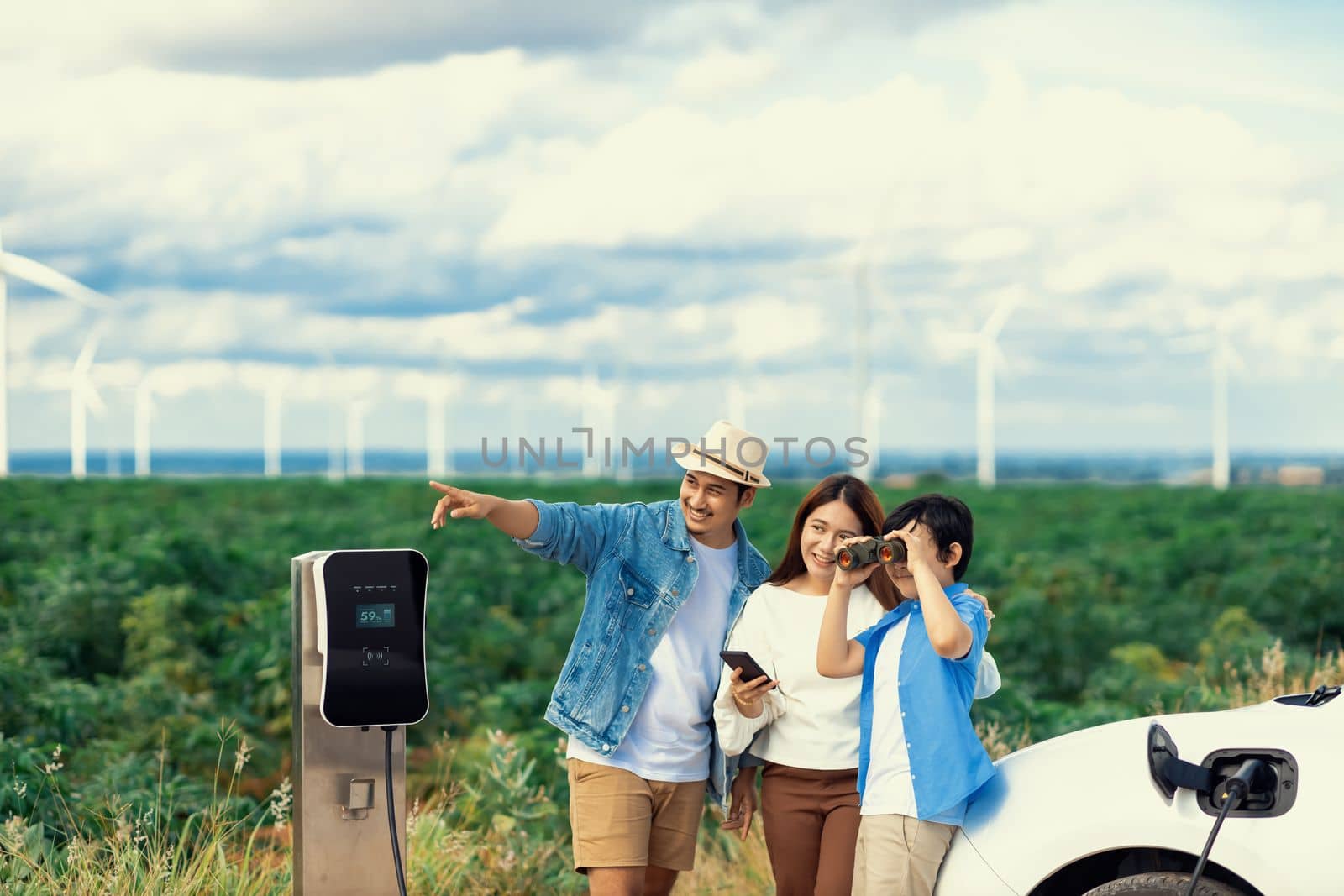 Concept of progressive happy family enjoying their time at wind farm with electric vehicle. Electric vehicle driven by clean renewable energy from wind turbine generator for charging station.