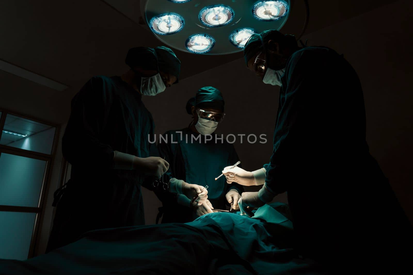 Surgical team performing surgery to patient in sterile operating room. In a surgery room lit by a lamp, a professional and confident surgical team provides medical care to an unconscious patient.