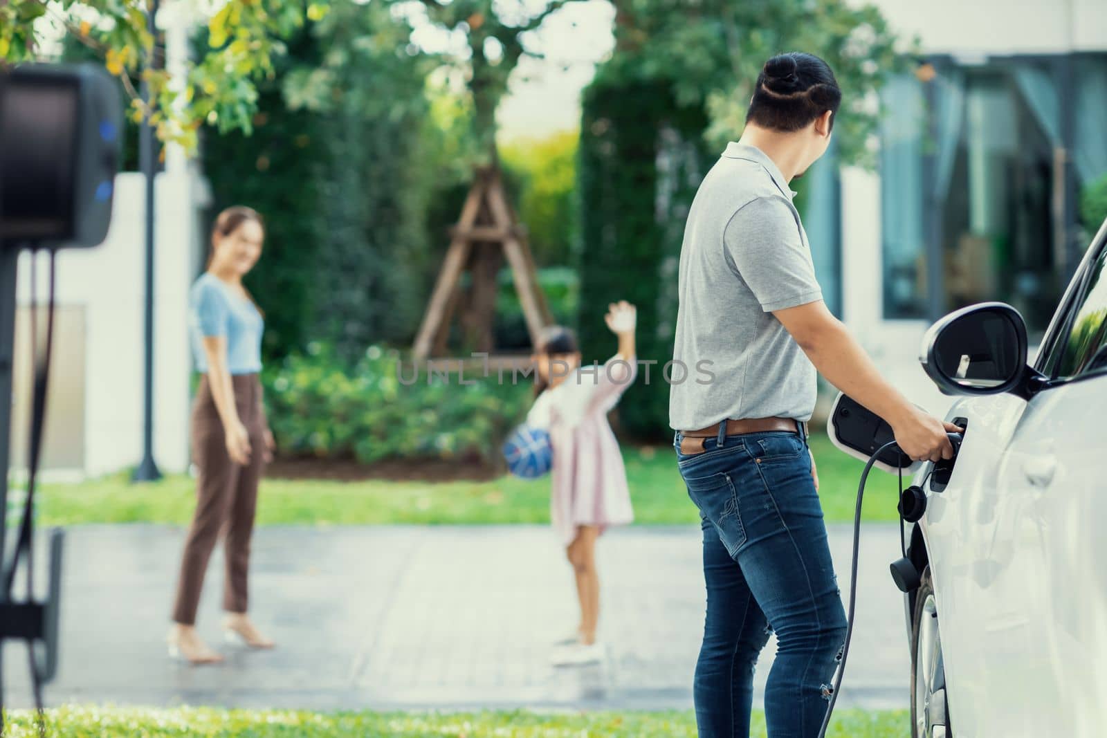 Focus image of progressive man charging electric car from home charging station with blur mother and daughter playing together in the background.
