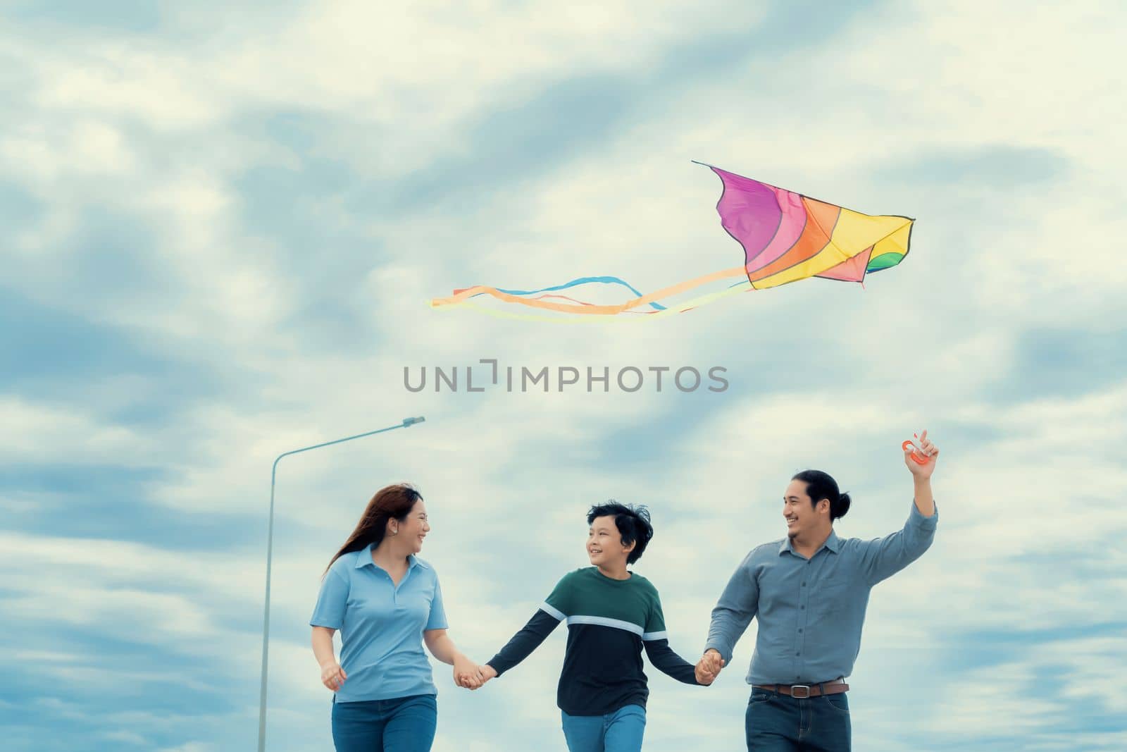 Progressive happy family vacation and carefree day concept. Young parents mother father and son run along and flying kite together road with enjoy natural scenic on scenery and clear sky background.