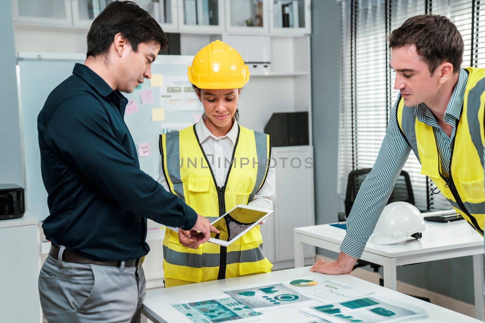 A team of competent engineers wearing safety equipment is working on blueprints with a tablet while also discussing with the investor in the office.