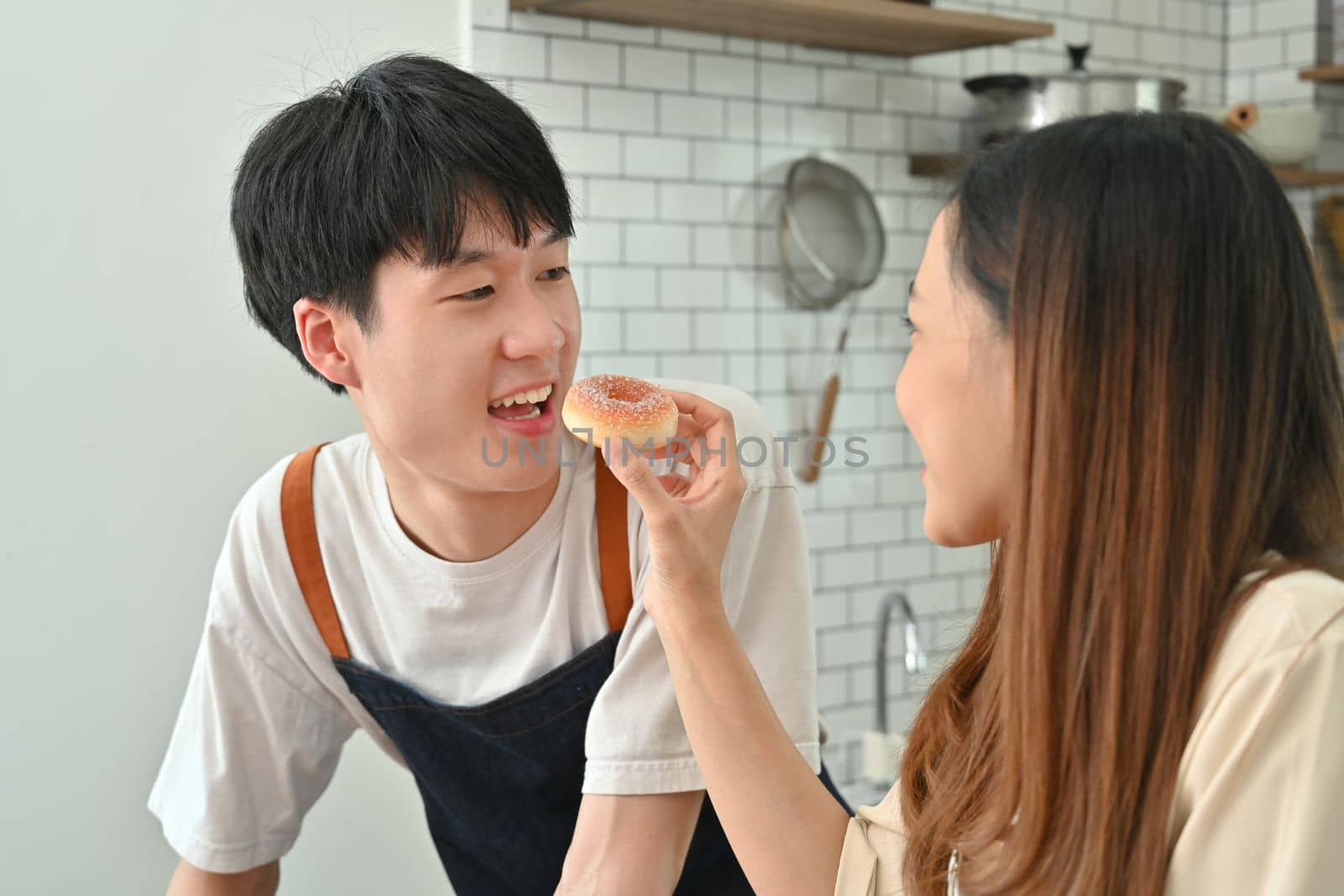 Young woman feeding her boyfriends with donut while cooking in modern at home, enjoying leisure weekend time together.