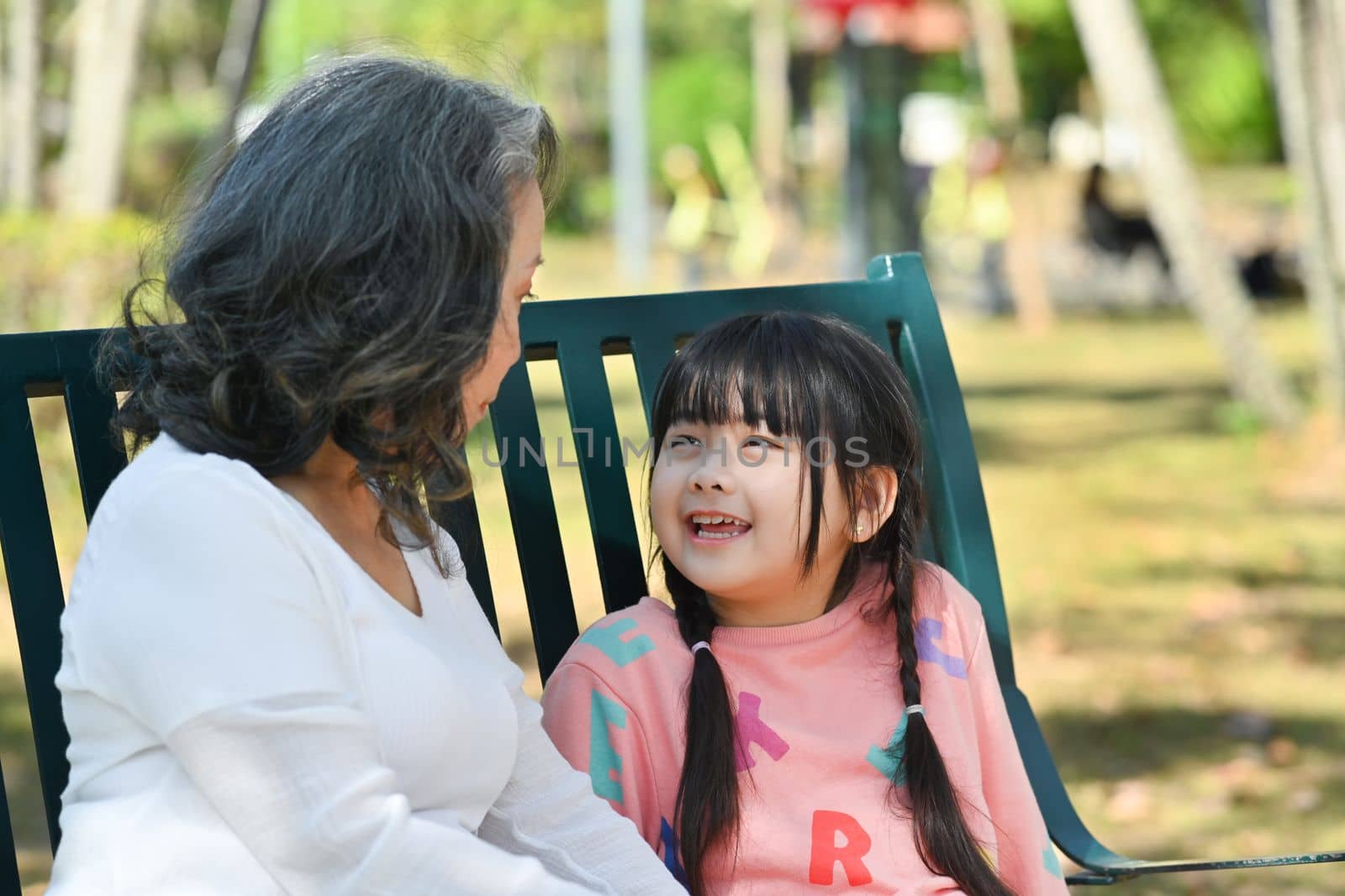Happy cute little grandchild enjoying talk with elderly grandmother while sitting on a bench in the park. Family and love concept by prathanchorruangsak
