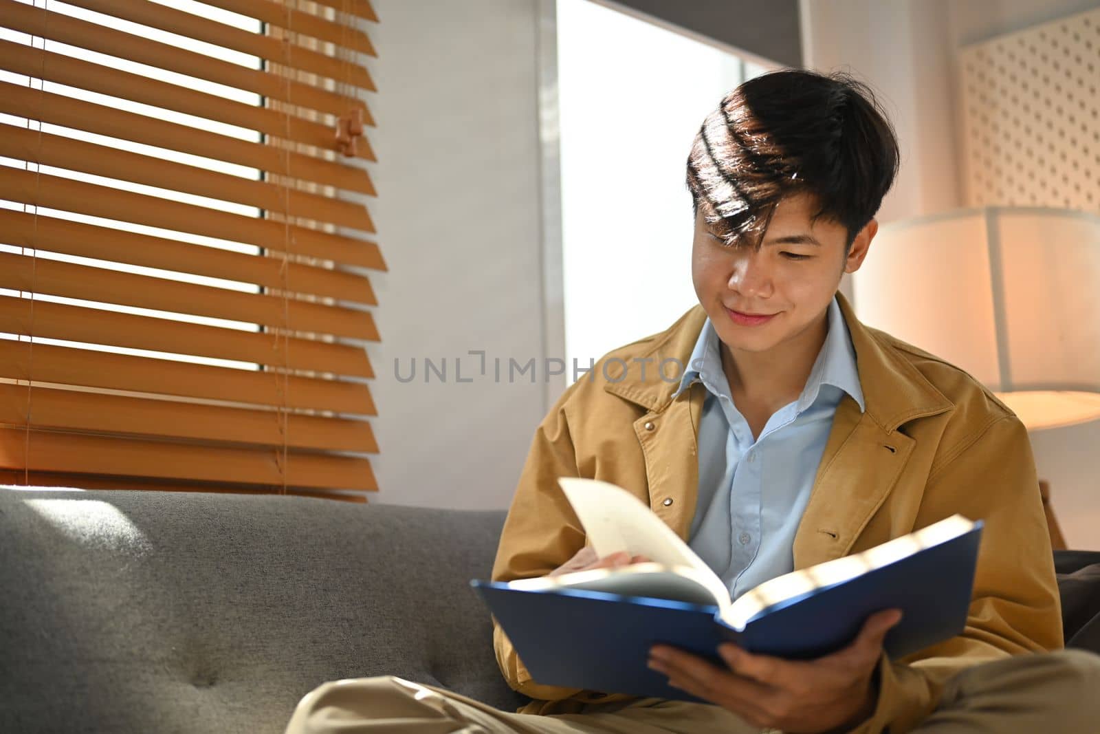 Relaxed millennial man reading book on comfortable couch, enjoying leisure weekend at home by prathanchorruangsak