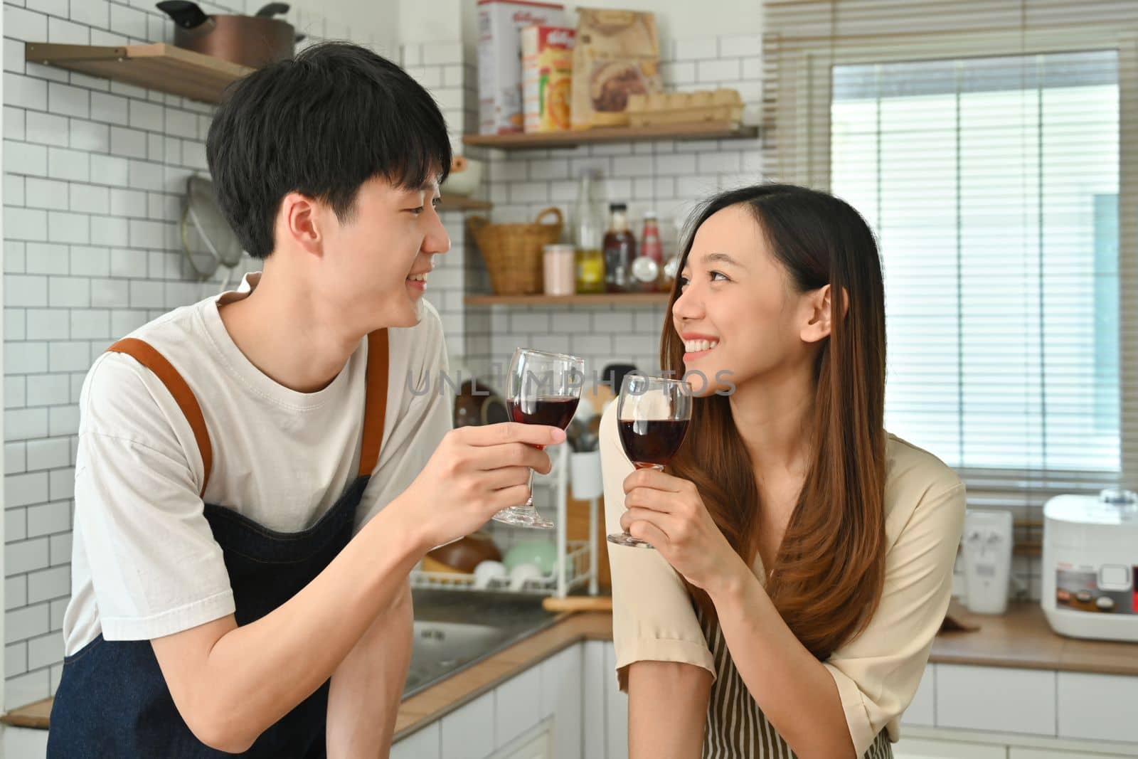 Romantic couple drinking red wine while preparing dinner in modern kitchen together.