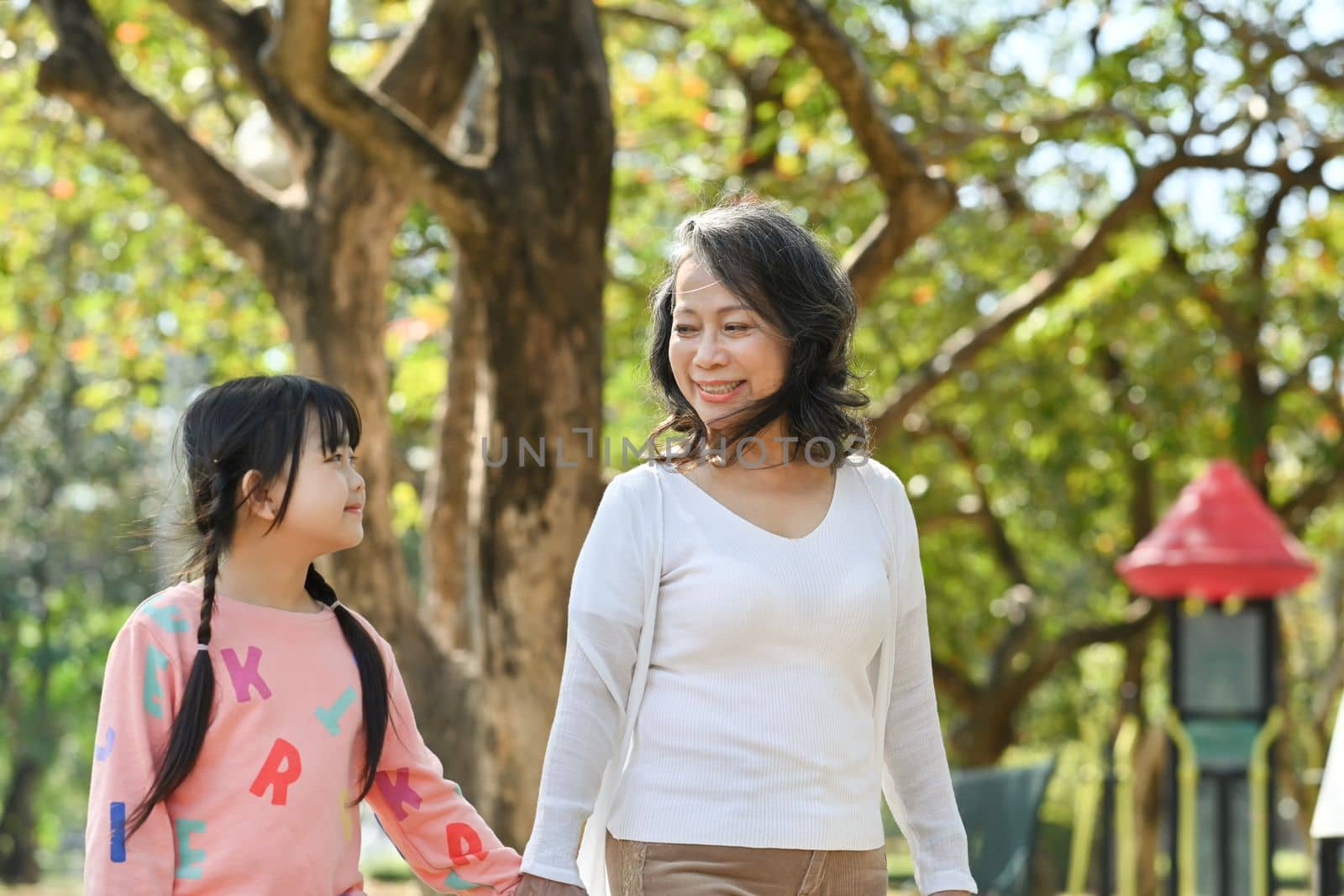Joyful little girl and grandmother walking in park surrounded by green trees at sunlight morning. Family, generation concept by prathanchorruangsak