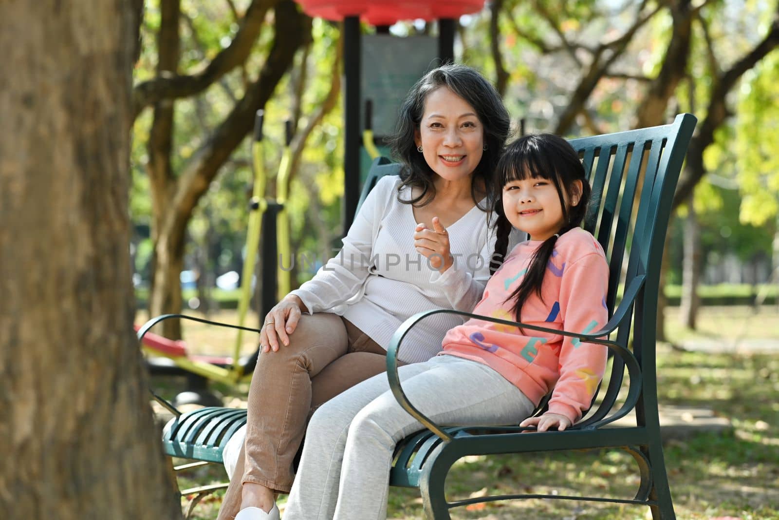 Photo of mature grandmother having fun enjoying talk with cute little grandchild while sitting on a bench in the park.
