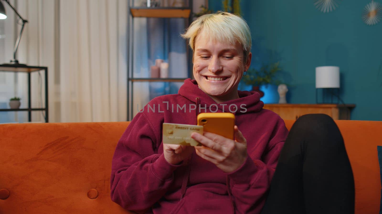 Young woman with short hair using credit bank card and smartphone while transferring money, purchases online shopping, order food delivery at home apartment indoors. Girl in evening room on couch