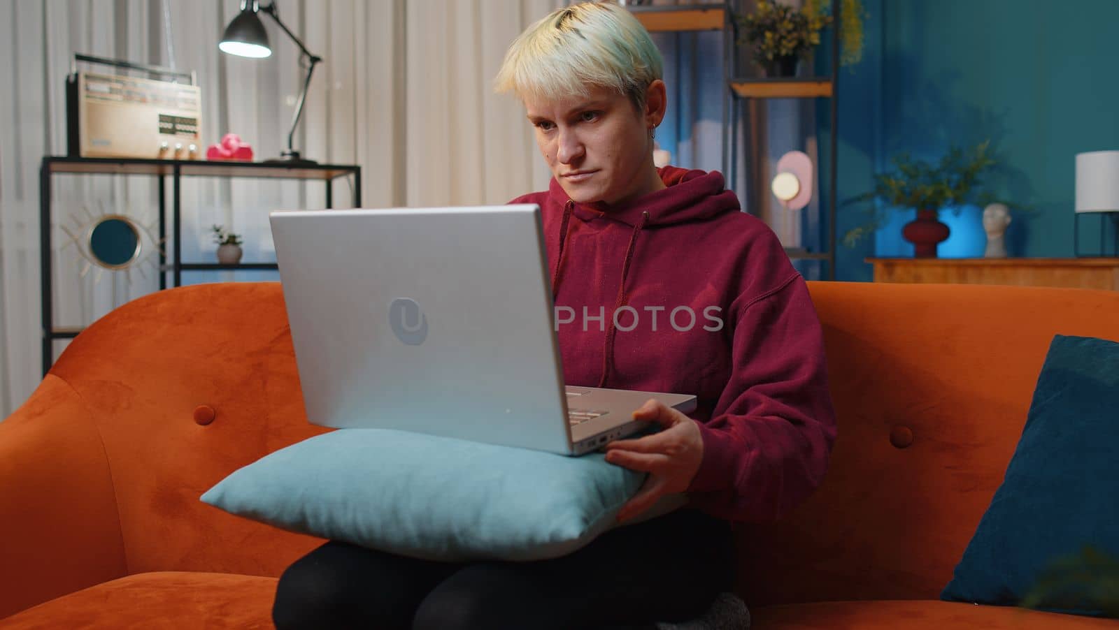 Young woman with short blonde hair freelancer at home night room sitting on couch, opens laptop start working. Girl works on notebook, sends messages, makes online purchases, watching movies, working