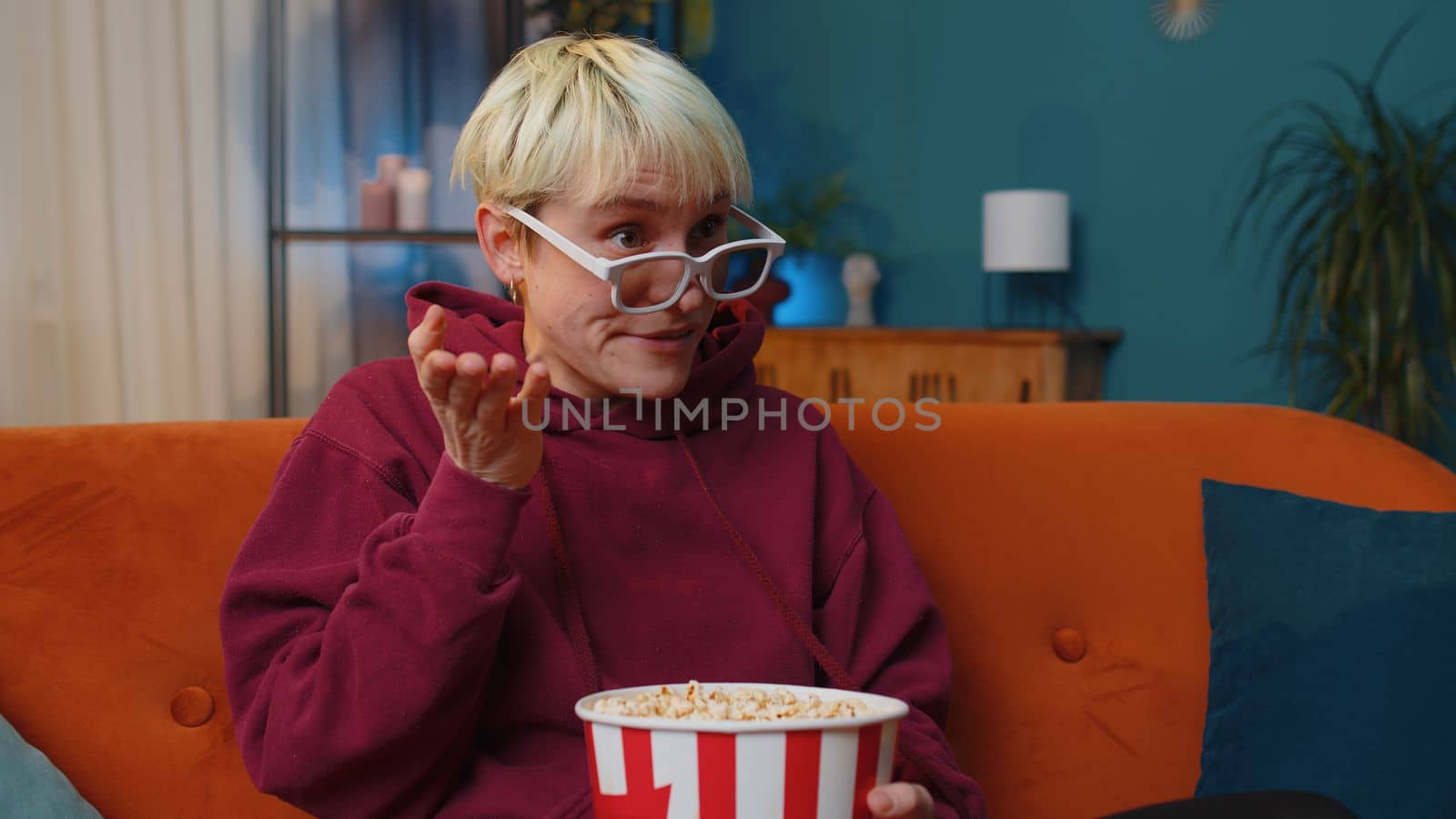 Excited young woman sits on sofa eating popcorn snacks and watching interesting TV serial, sport game, film, online social media movie content at home. Girl enjoying evening entertainment. Lifestyles