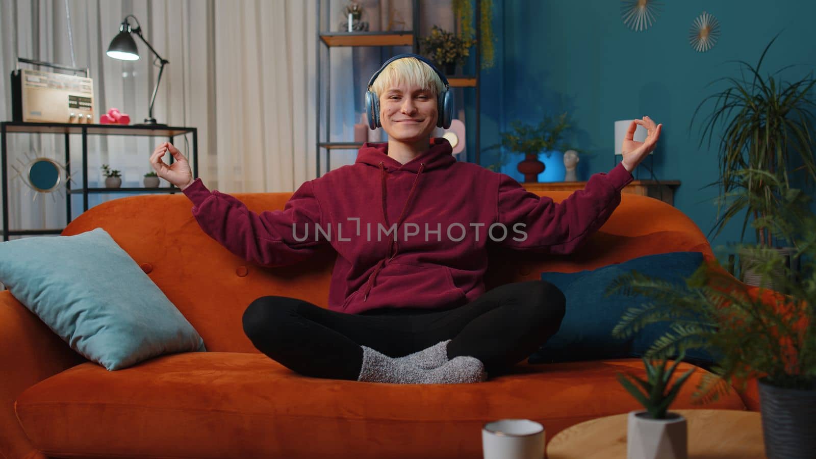 Keep calm down, relax. Woman with short hair at home couch breathes deeply with mudra gesture, eyes closed meditating with concentrated thoughts, peaceful mind, listen music. Girl in evening room