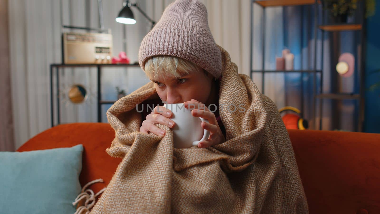 Sick young woman wrapped in plaid sitting, shivering from cold on sofa drinking hot tea in unheated apartment without heating due debt. Unhealthy girl feeling discomfort try to warm up at evening home