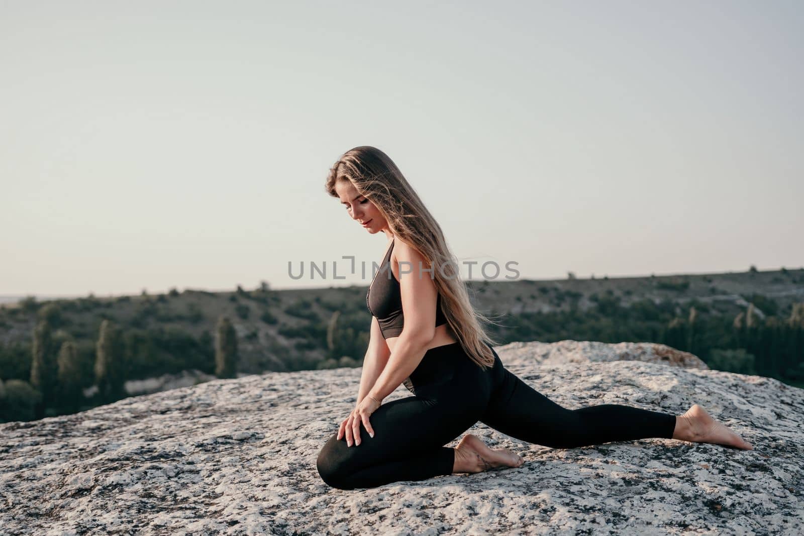 Well looking middle aged woman with long hair, fitness instructor in leggings and tops doing stretching and pilates on the rock near forest. Female fitness yoga routine concept. Healthy lifestyle.