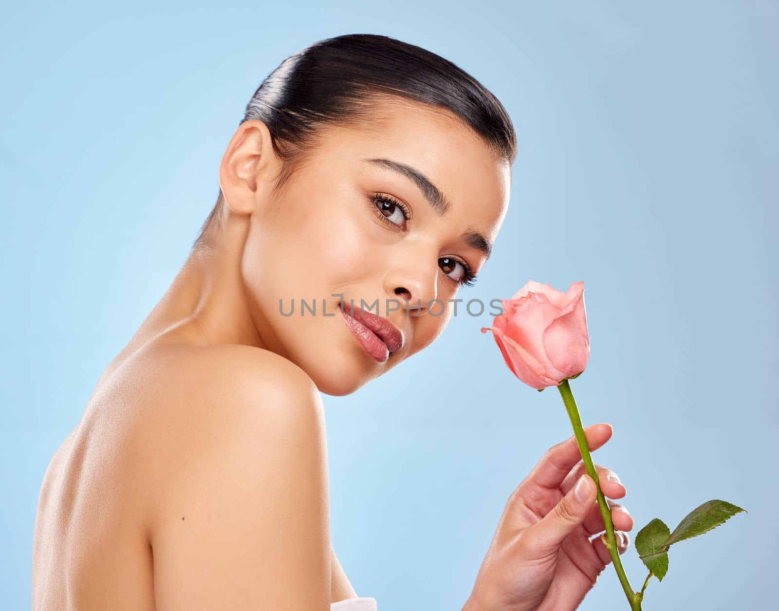 Her natural beauty blooms like no other. Studio portrait of an attractive young woman posing with a pink rose against a blue background. by YuriArcurs