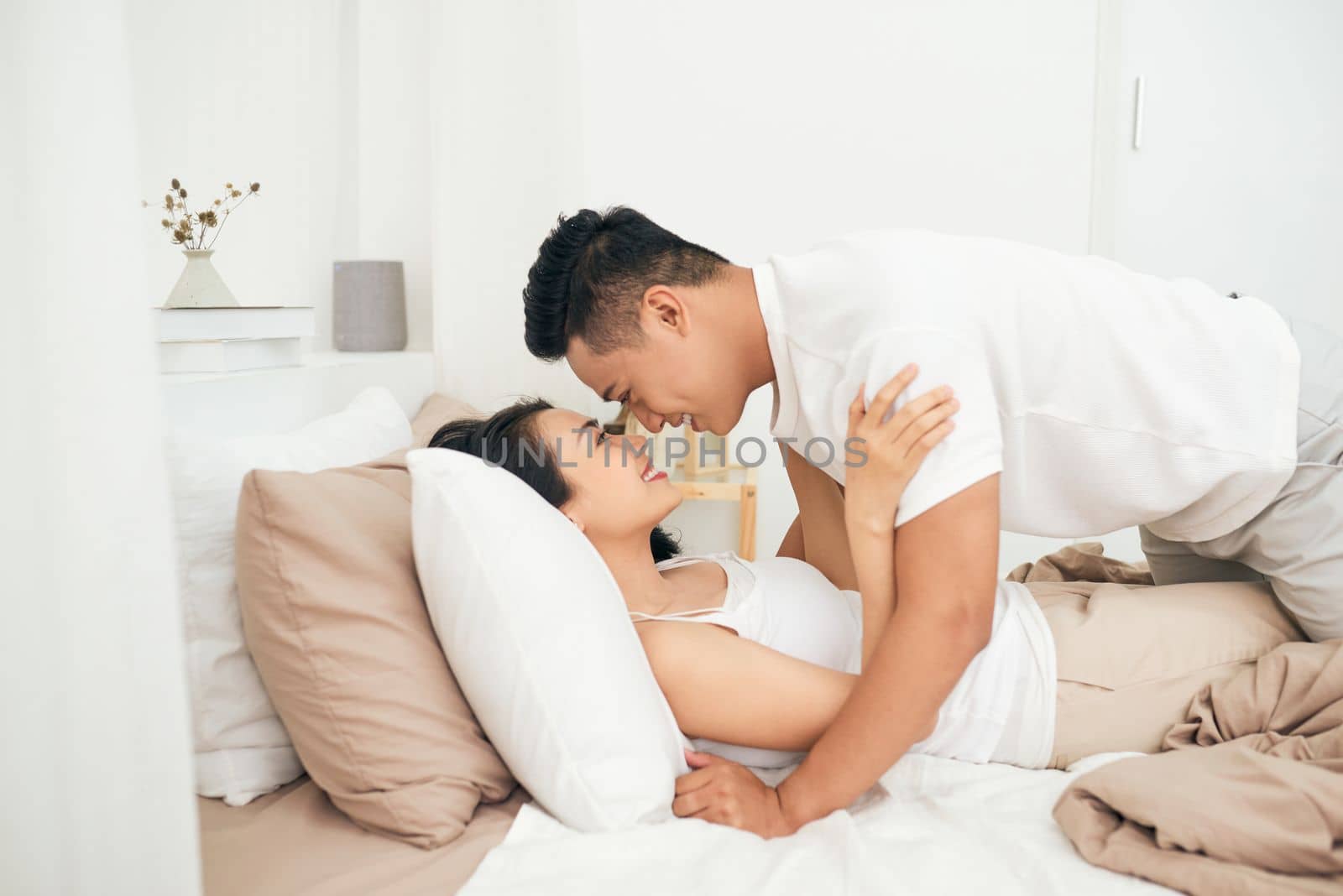 Side view of excited man is lying over woman in bed. They are looking at each other eyes and laughing