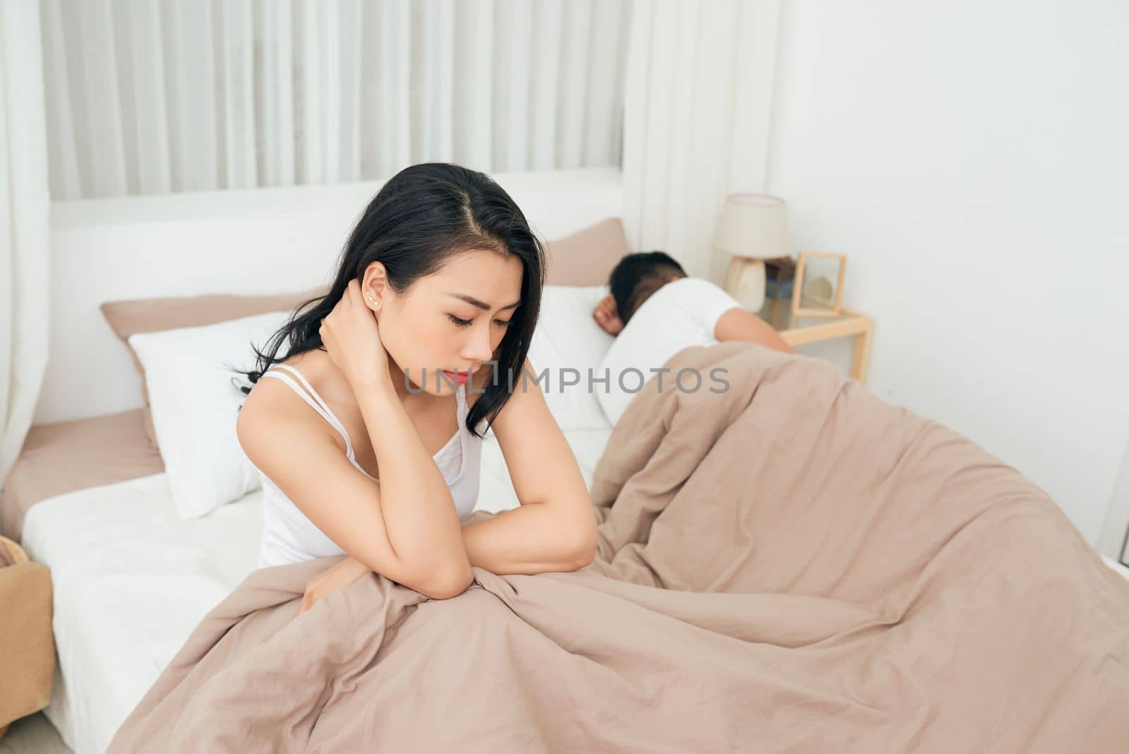 We have a problem. Young upset girl sitting on the edge of the bed, against her boyfriend, lying in bed.