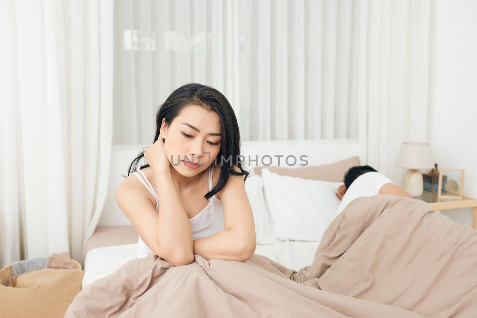 We have a problem. Young upset girl sitting on the edge of the bed, against her boyfriend, lying in bed.