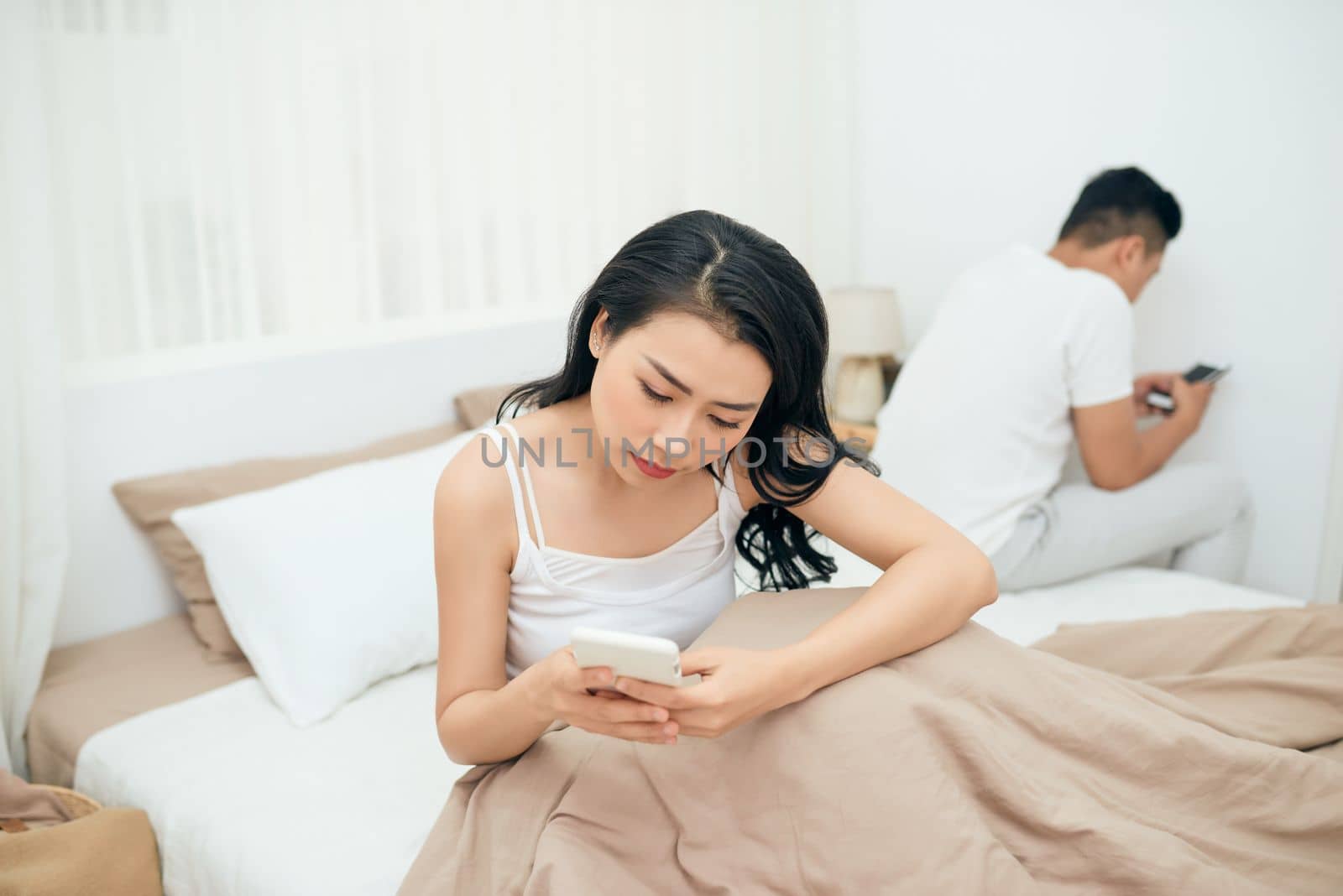 sad view of young married couple using their mobile phone in bed ignoring each other as strangers in relationship and communication problems by makidotvn