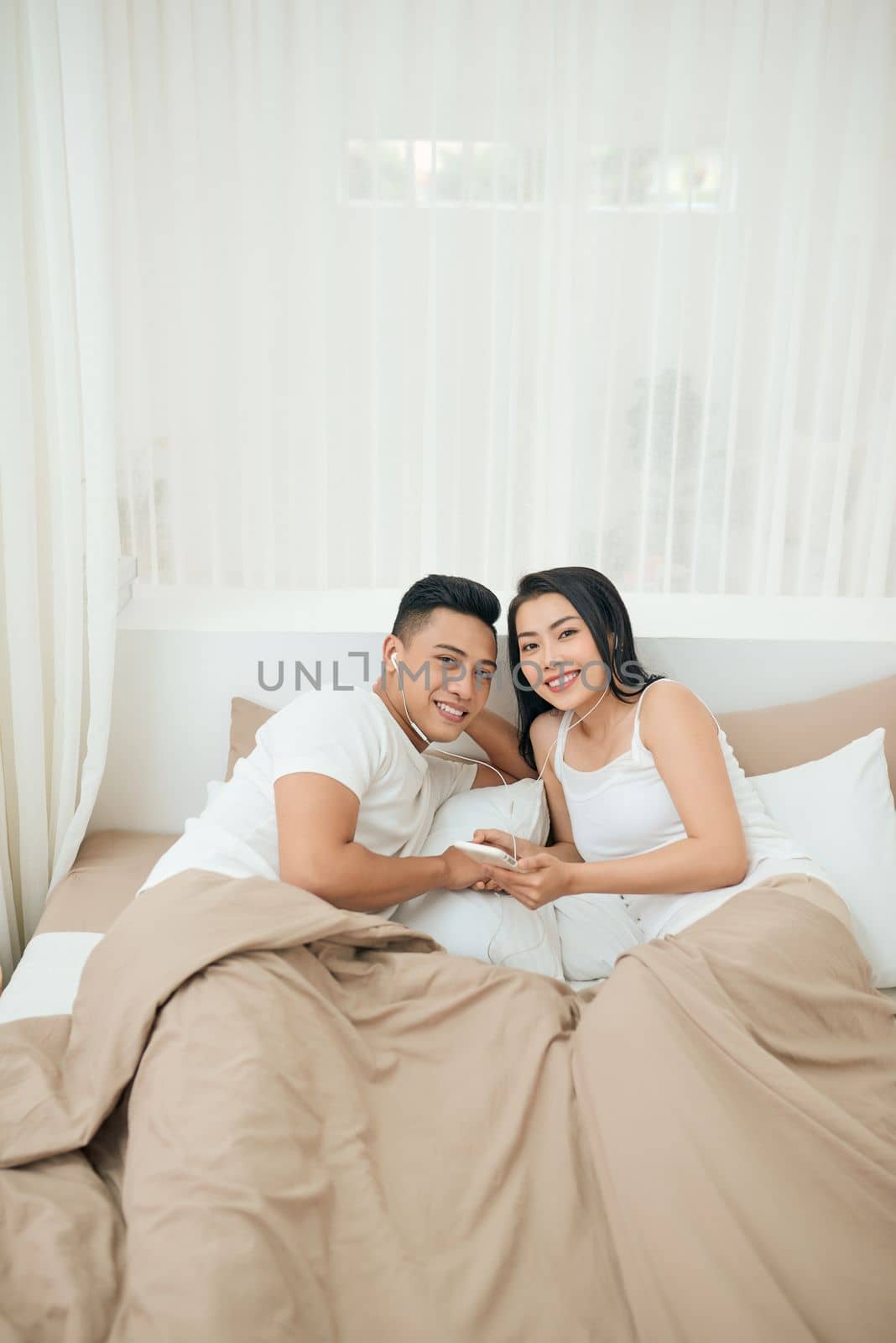 Young asian couple smile listen music with smart mobile phone on bed at bedroom with fun and enjoy